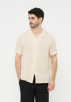 Mężczyzna o ciemnych włosach, ubrany w kremową, krótkawą, zapinaną na guziki koszulę oraz czarne spodnie, patrzy w lewo ze schowanymi w kieszeniach rękami.