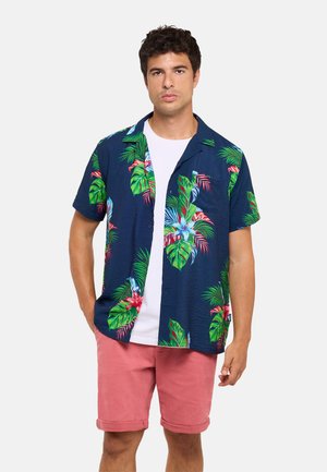 Hombre con una camisa de manga corta azul marino con estampado floral tropical sobre una camiseta blanca y pantalones cortos rosas, posando frente a un fondo liso.