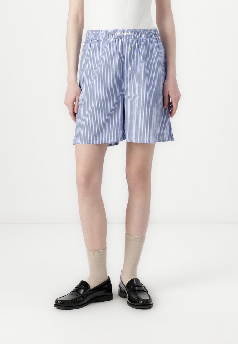 Modelka w niebiesko-białych pionowo prążkowanych szortach z elastyczną talią, beżowych skarpetkach i czarnych skórzanych mokasynach na białym tle.