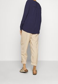 Haut bleu marine à manches longues en tissu texturé avec un dos plissé, associé à un pantalon beige retroussé et des chaussures légères à enfiler de couleur claire.