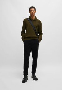Suéter de punto oversize en caqui con cuello alto, combinado con pantalones de pana negros y botas negras con cordones, que incluyen una correa de hombro.