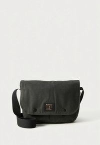 Borsa a tracolla in tela verde con chiusura a patta, tracolla nera e etichetta marchiata. Texture liscia, dettagli minimalisti e design pratico.