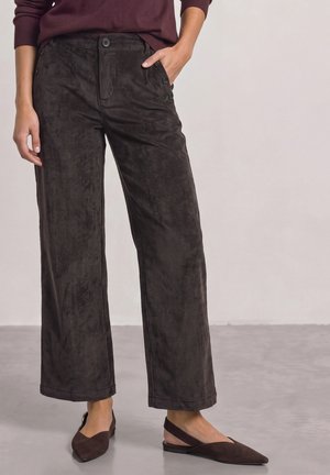Personne portant un pantalon en velours côtelé marron foncé à jambes larges et des ballerines marron à bride arrière assorties, debout sur un sol gris.