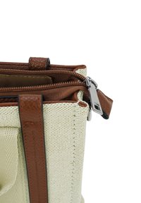 Nahaufnahme einer beigen, strukturierten Stofftasche mit braunen Lederträgern und teilweise geöffnetem Reißverschluss, der die obere Kante und den Metallreißverschluss zieht.