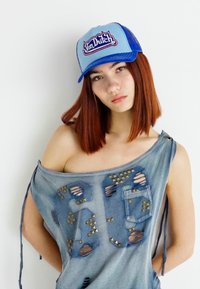 Casquette de camionneur bleu et gris avec le logo "Von Dutch" ; haut en denim déchiré avec des découpes et des clous métalliques ; coupe décontractée et design épaules nues.