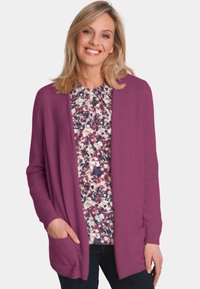 Langärmlige, offene Strickjacke in kräftigem Lila, mit zwei seitlichen Taschen und gerippten Bündchen, über einer blumengemusterten Bluse getragen.