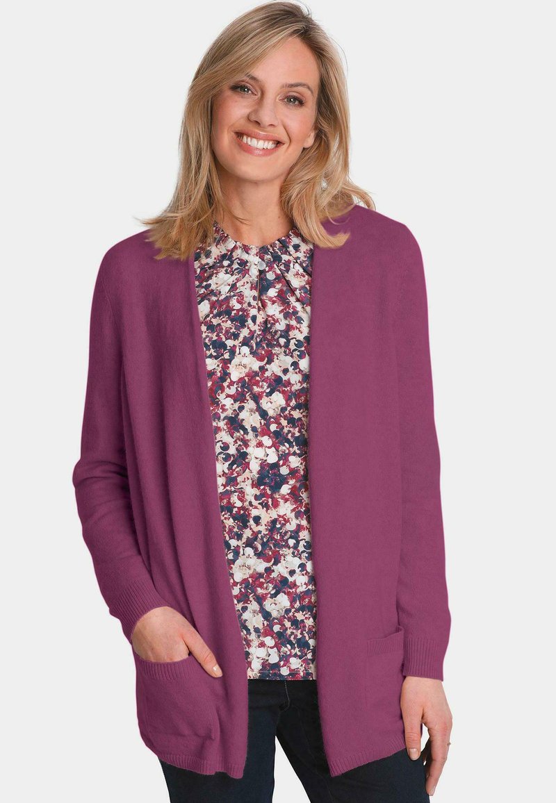 Langärmlige, offene Strickjacke in kräftigem Lila, mit zwei seitlichen Taschen und gerippten Bündchen, über einer blumengemusterten Bluse getragen.