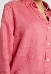 Chemise en lin rose avec un devant à boutons, un col et des manches longues. Texture douce avec un tissage de tissu visible. Présente des boutons blancs le long de la patte de boutonnage.