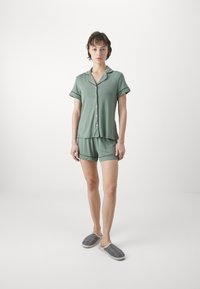 Zeleni kratki pajama set s kratkimi rokavi in gumbi, z zavihanimi manšetami in ujemajočimi kratkimi hlačami. Pohan z sivimi kockastimi copati.