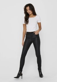 Vit kortärmad topp kombinerad med åtsittande svarta byxor i konstläder. Modellen bär svarta högklackade skor. Enkel, elegant design.