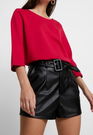 Woman wearing a red three-quarter sleeve top tucked into high-waisted black leather shorts with a wide black belt with a square buckle.