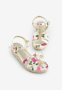 Białe kwiatowe sandały na koturnie z różowymi różami, z grubym obcasem, regulowanym paskiem w kostce i metalicznym akcentem na przednim kokardzie.