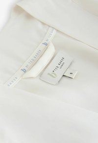 Material alb cu o textură netedă, având o etichetă inscripționată "b by Ted Baker" și o etichetă de mărime "S" în verde și negru.