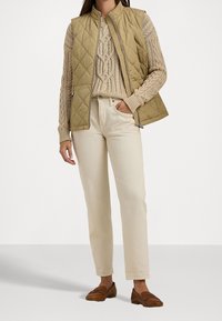 Tan quiltad väst bärs över en krämfärgad kabelstickad tröja, i kombination med ljusa byxor och bruna loafers. Enkel, funktionell design med vertikala mönster.