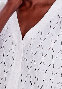 Haut en tissu texturé blanc avec un décolleté en V, présentant des découpes décoratives et une fermeture à boutons sur le devant. Design léger et détaillé.