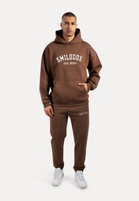 Brązowa bluza z kapturem z napisem "SMILODOX ATHL. WEAR" na białym tle, w komplecie z dopasowanymi brązowymi spodniami dresowymi. Teksturowany materiał, kieszeń kangurka, białe sneakersy.
