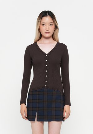 Brown ribbed long-sleeve cardigan with a lace-trimmed V-neck and pearl buttons, paired with a navy plaid mini skirt featuring a side slit.