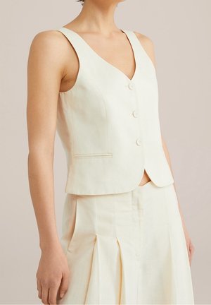 Femme portant un gilet crème sans manches boutonné et une jupe plissée assortie sur un fond uni.