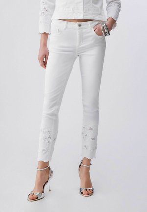 Žena v bílých úzkých kalhotách s květinovými krajkovými výřezy u kotníků, bílém topu, stříbrných náramcích a stříbrných sandálech na vysokém podpatku.