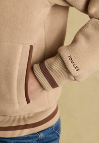 Beezhe fliisjakiga, ribidega mansettide ja äärmusega, millel on pruunid triibud, käsi küljetaskus ja "Joules" tikand mansetil.