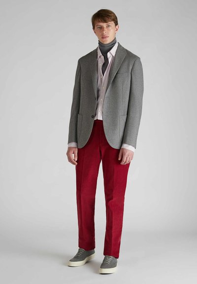 Grau maßgeschneiderter Blazer über einem gestreiften Hemd, kombiniert mit roten Cordhosen und grauen Sneakers, steht vor einem neutralen Hintergrund.
