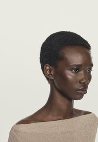 Vista laterale di una persona con capelli corti e ricci che indossa un top beige textured scollato, che guarda in avanti contro uno sfondo neutro.