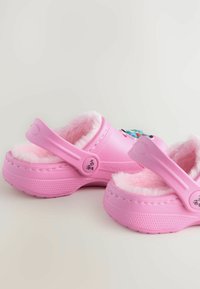 Roze klompen met een zachte, fluffy voering, verstelbare band en ronde decoratieve accenten. De zijkant heeft textuur met perforaties voor ventilatie.