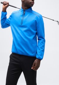 Άνδρας με φωτεινό μπλε πουλόβερ Calvin Klein κρατά μια ράβδο γκολφ που ακουμπά στον ώμο του, ντυμένος με μαύρο παντελόνι σε απλό φόντο.