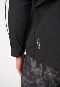 Veste légère noire avec une finition texturée, présentant un logo "ADIZERO" vert fluo sur le côté, associée à un short gris à motifs.