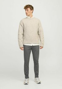 Suéter de punto beige con cuello redondo, combinado con pantalones ajustados gris oscuro. El modelo lleva zapatillas grises y blancas, presentando un atuendo casual.