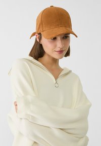 Casquette de baseball en cuir suédé brun avec une visière incurvée, des détails de couture et une sangle arrière ajustable, portée avec un pull zippé crème clair.