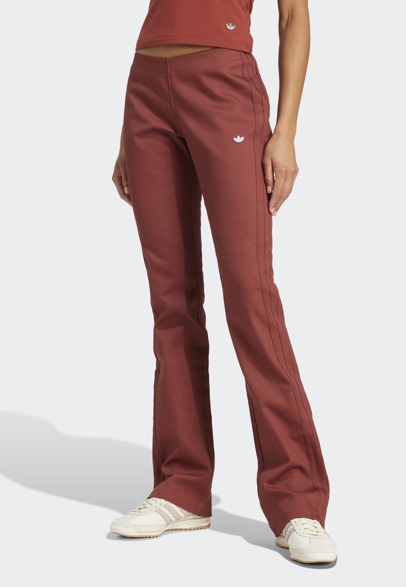 Personne portant un pantalon évasé couleur rouille avec le logo Adidas, des baskets blanches et un haut court assorti sur un fond uni.