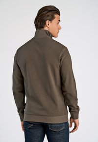 Brown, high-collar sweatshirt with long sleeves, textured fabric, ribbed cuffs, and a straight hem, paired with dark blue jeans.