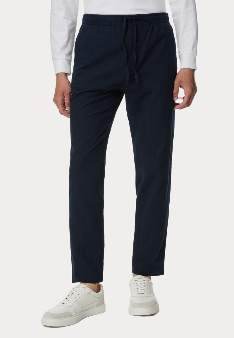 Pantaloni da jogging in blu navy, tessuto intrecciato con vita elastica e cordino. Tessuto testurizzato, aderenti alla caviglia. Abbinati a sneakers bianche.