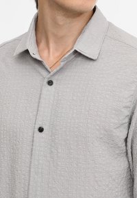 Camisa de botones texturizada en gris claro, con cuello y botones negros. Presenta un patrón sutil en toda la tela.