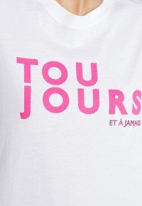 Bílý bavlněný tričko s výrazným růžovým nápisem "TOUJOURS" a menším textem "ET À JAMAIS", ozdobený kamínky.