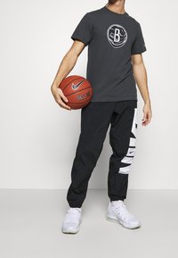 Pantalons de sport noirs avec logo Nike blanc, t-shirt gris avec graphisme de basketball, tenant un ballon de basketball Nike rouge, baskets blanches.