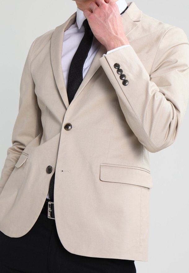 Blazer beige hecho de tela suave, cierre de un botón, solapas con muesca y dos bolsillos frontales. Combina con una corbata negra y una camisa blanca.
