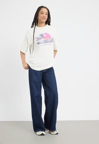T-shirt blanc présentant un graphisme "La Grande Vague" en rose et bleu, associé à un jean en denim foncé à jambes larges et des chaussures de sport.