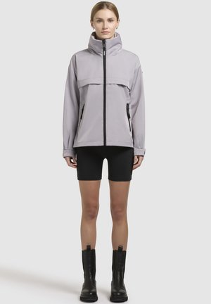 Veste imperméable gris clair avec col haut, fermeture éclair frontale et poches latérales. Associée à un short noir et des bottes noires hauteur cheville.