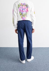 Camicia bianca a maniche lunghe con grafica colorata e testo sul retro; pantaloni blu navy con dettaglio della tasca posteriore; indossati con sneakers bianche.