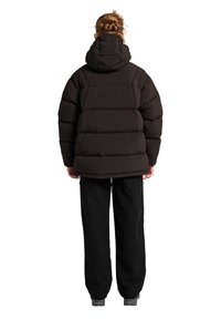 Kobieta z zaplecionymi włosami, ubrana w czarną puchową kurtkę z kapturem, szerokie czarne spodnie i szare buty, stoi tyłem.