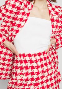 Röd och vit rutad blazer- och kjolset i texterat tyg, kombinerat med en enkel vit off-shoulder topp. Gyllene knappdetaljer synliga.