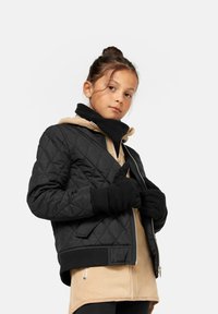 Svart quilted jakke med ribbestrikket nederkant, kombinert med en beige, zip-up hettejakke. Modellen har på seg svarte hansker og står mot en hvit bakgrunn.