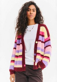 Kolorowy sweter na guziki z pionowymi paskami w odcieniach różu, fioletu, żółci i bordowego. Posiada cztery guziki oraz miękką, fakturowaną tkaninę.