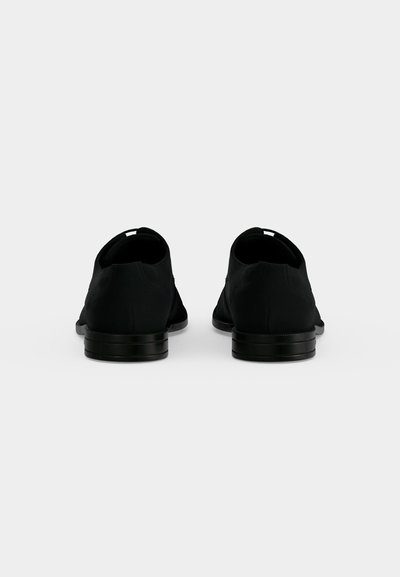 Paire de chaussures noires formelles vues de dos, montrant des talons bas et des dessus en cuir lisse sur un fond clair uni.