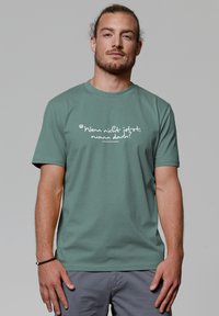 Mann mit leichtem Bart und zurückgebundenem Haar trägt ein grünes T-Shirt mit dem weißen deutschen Text "Wenn nicht jetzt, wann dann?" vor einem einfarbigen Hintergrund.