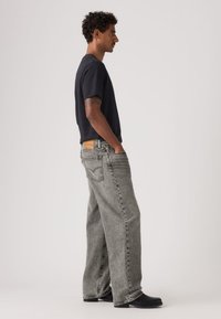 Γκρι τζιν άνετης εφαρμογής με δερμάτινο ταμπελάκι στη μέση, συνδυασμένα με μαύρο κοντομάνικο μπλουζάκι και μαύρα παπούτσια. Εμφανίζεται από πλάγια όψη.