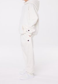 Barn som bär en vit Champion-hoodie, matchande sweatsbyxor och vita sneakers, står på sidan med händerna i fickorna mot en vit bakgrund.