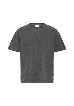 Ung mand med kort hår iført en vasket sort oversized t-shirt, sorte bukser og sorte sko, der står mod en lys baggrund.